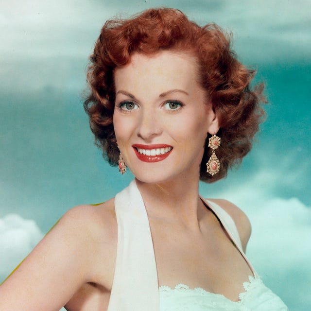 maureen o'hara smiles at the camera, she wears red lipstick, large dangling earrings and a white halter top