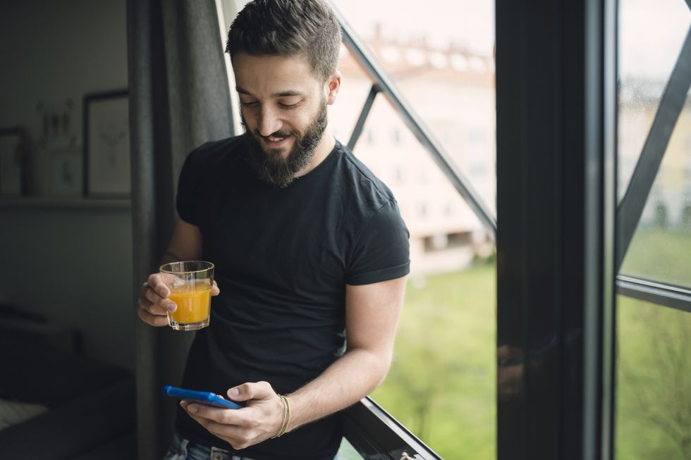 Young man drinking an orange juice and using his smartphone at the window