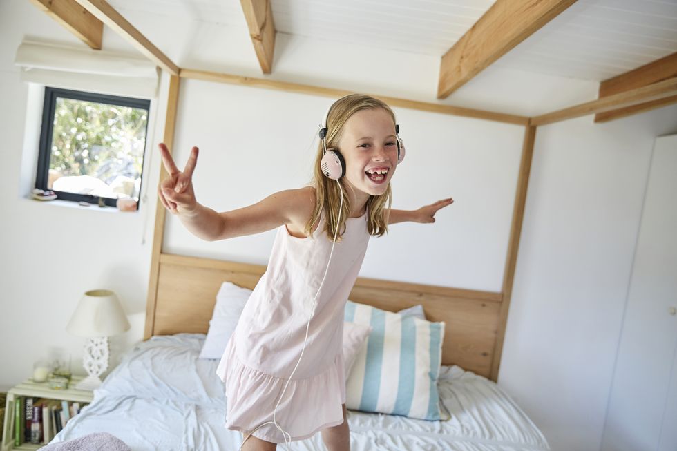 Portrait of smiling little girl dancing on bed at home while listening music with headphones