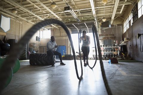 Male personal trainer watching female client doing gym battling ropes exercise in gym
