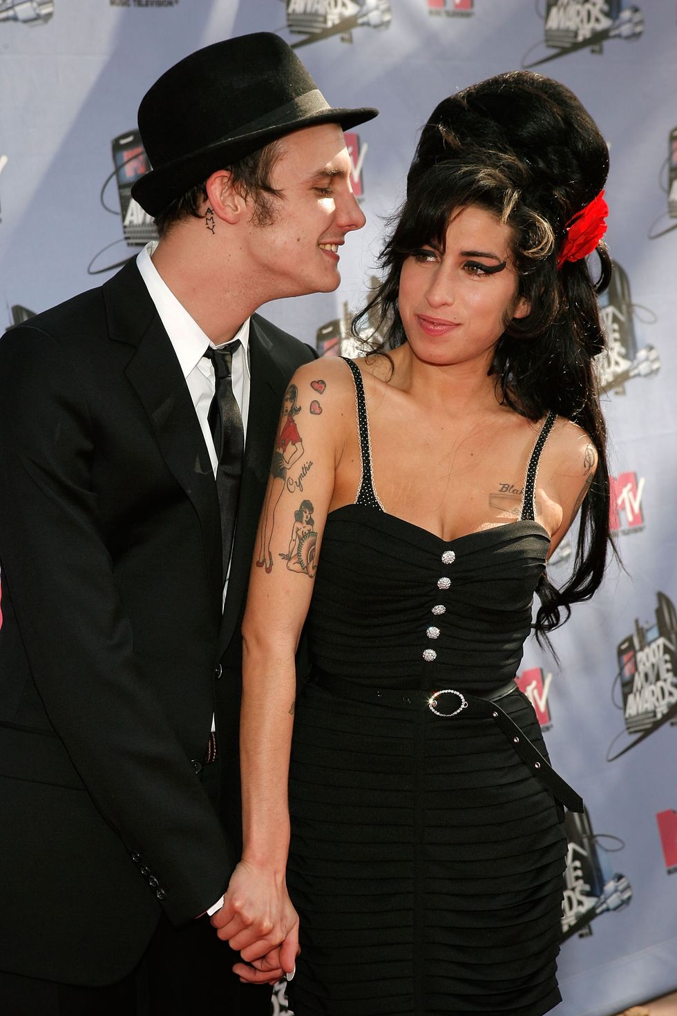 blake fielder civil and amy winehouse stand together in front of lavender photo backdrop with logos, he looks at her and smiles and she looks to the left and smiles, they hold hands and both wear black outfits