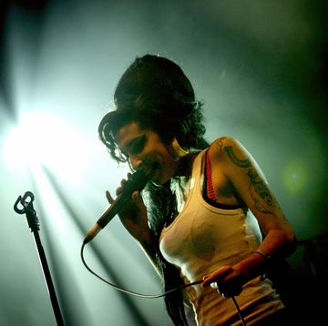 amy winehouse sings into a microphone she holds in one hand, she smiles and stands next to a microphone stand, she wears a white tank top