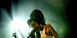 amy winehouse sings into a microphone she holds in one hand, she smiles and stands next to a microphone stand, she wears a white tank top