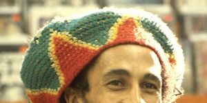bob marley smiles and wears a red, yellow and green knit hat with a denim collared shirt over an orange v neck sweater