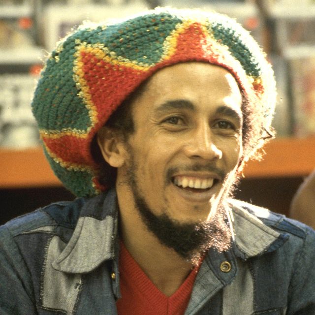bob marley smiles and wears a red, yellow and green knit hat with a denim collared shirt over an orange v neck sweater