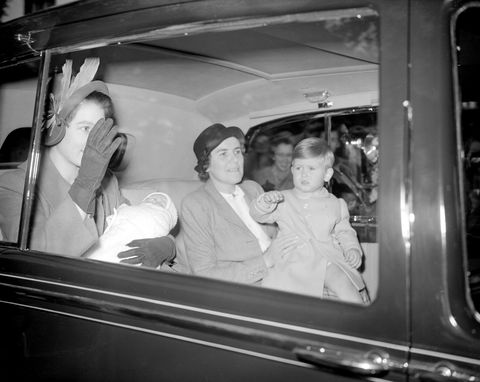 princess elizabeth waves while holding princess anne as an infant, a young prince charles also waves while sitting on a womans lap, all four people are riding inside the backseat of a car