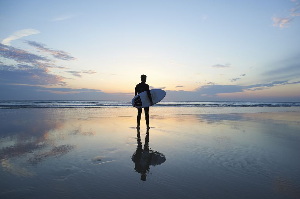 a surfer standing on a deserted beach with a beautiful sunset reflected in the shallow waters