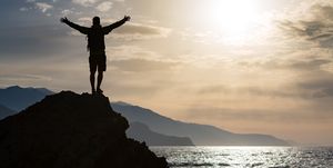 man with arms outstretched celebrating or praying in beautiful inspiring sunrise with mountains and sea man hiking or climbing with hands up enjoy inspirational landscape on rocky top on crete