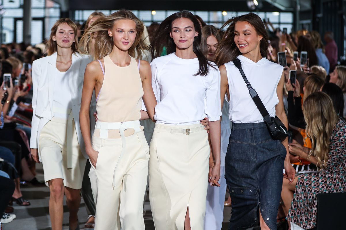 three modern, minimalist models walk down the runway for tibi's spring 2018 show a blonde woman wears a cream tank top with paler cream low slung pants a brunette wears a white tee with a pale cream skirt and another brunette wears a white sleeveless top with a denim skirt