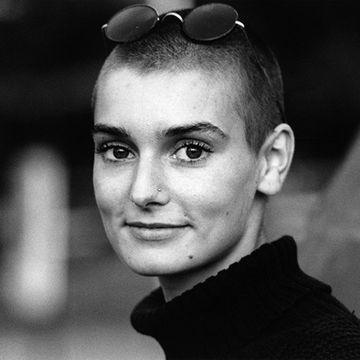 sinead o'connor smiles at the camera, she wears a turtleneck sweater and glasses on her head
