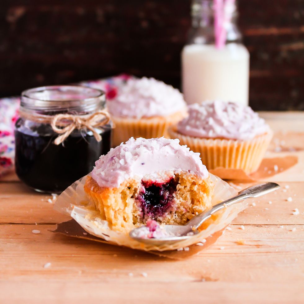 Homemade vanilla cupcakes with berry jam, coconut and cream cheese with a jar of jam and a bottle of milk on a wooden table, selective focus