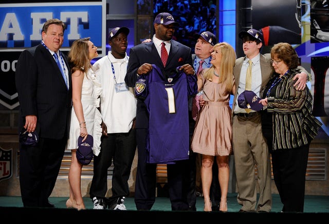 michael oher stands in a group of people while holding up a baltimore ravens jersey, he wears a suit and a baltimore ravens hat, his family are dressed in business formal clothes