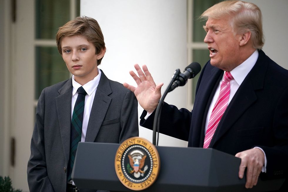 barron trump stands to the side of his father who speaks into a microphone on a podium with a presidential seal, both wear suit jackets with white collared shirts and ties