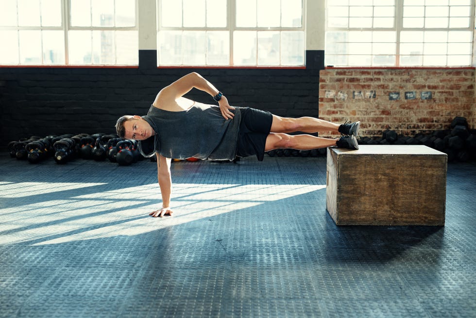 shot of a young man doing a side plank with a wooden block in a gym