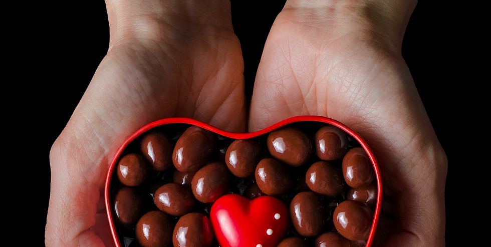 human hands holding red heart shaped box full of chocolate