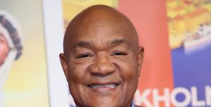 george foreman smiles at the camera, he wears a tan plaid suit jacket with blue and white accents and a blue chambray collared shirt