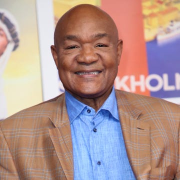 george foreman smiles at the camera, he wears a tan plaid suit jacket with blue and white accents and a blue chambray collared shirt