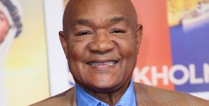 george foreman smiles at the camera, he wears a tan plaid suit jacket with blue and white accents and a blue chambray collared shirt