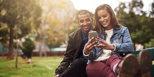 shot of a cheerful young couple sitting down on a bench while using a phone together outside in a park