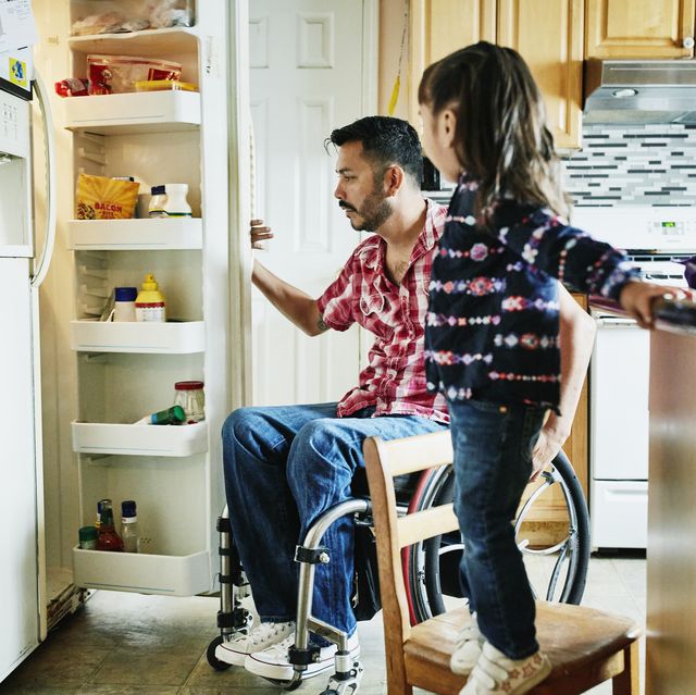Father in wheelchair looking in refrigerator in kitchen while daughter stands on chair watching