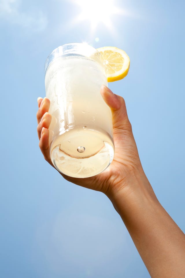 asian woman holding up a glass of lemonade with a slice of lemon on the edge of the glass with blue sky and sunburst in the background 