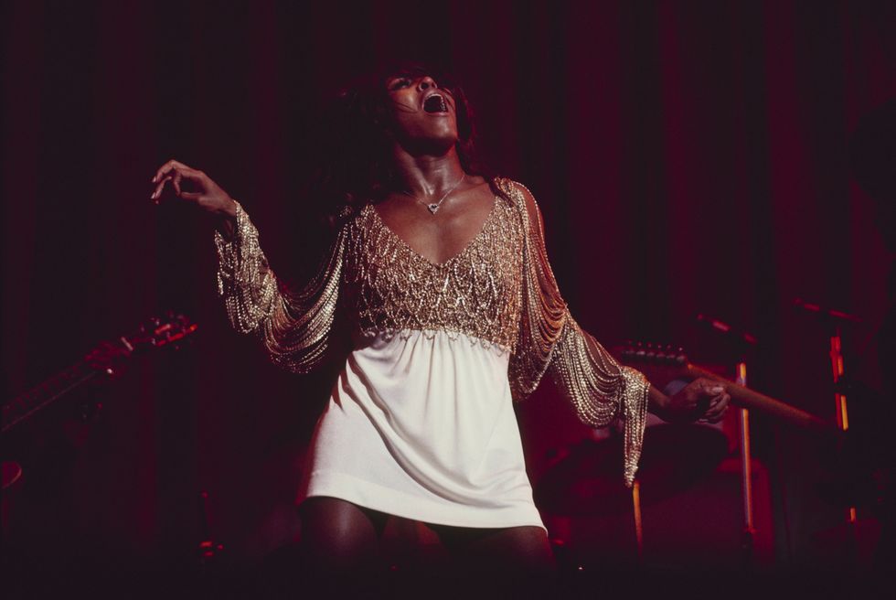 tina turner performs on stage, her head is lifted upward and her mouth open while singing, she is wearing a white skirt with a fringe top, her arms are extended at her sides