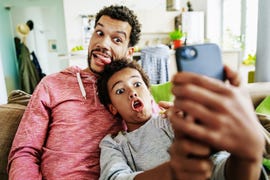 a father and son at home pulling silly faces while taking a selfie together on a smartphone