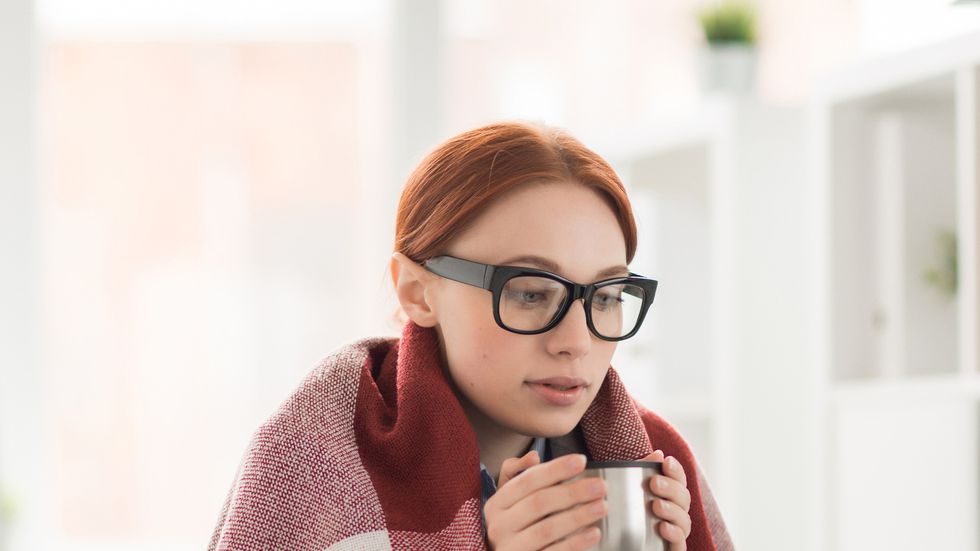 young woman in plaid and eyeglasses drinking hot tea from mug during work in office