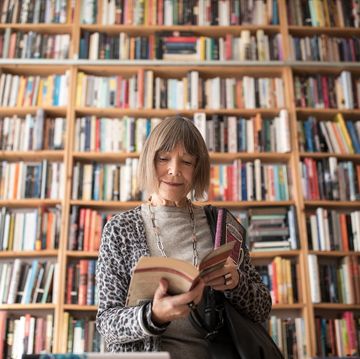 senior woman reading book while standing against bookshelf at bookstore