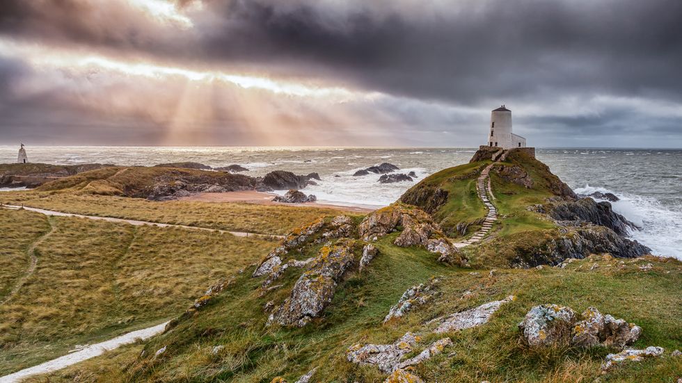 twr mawr and twr bach on ynys llanddwyn, anglesey, wales, uk
december 21, 2014 
winter view of llanddwyn island lighthouse the lighthouse sits high on the cliff with a narrow pathway of steps leading up to the door storm clouds and the sun rays beam down out to sea whilst large waves crash against the rocky beach below