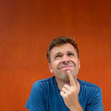 attractive thoughtful young man is confused and unsure he is sitting near red wall