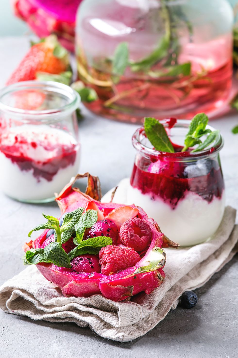 vegan fruit salad with berries and mint served in pink dragon fruit on cloth with jars of yogurt, bottle of lemonade, ingredients above on grey table close up healthy eating