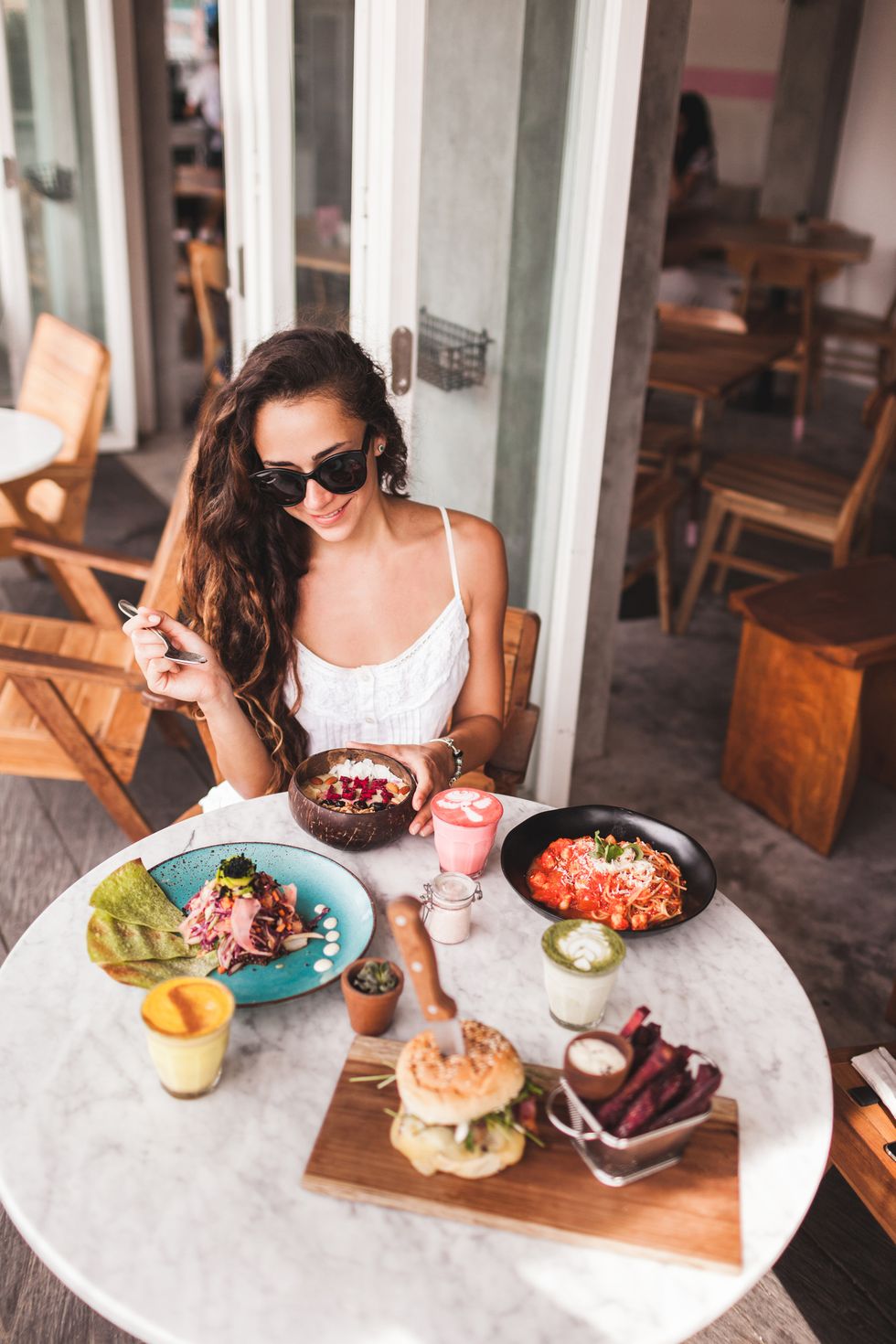 young smiling woman having breakfast in stylish street cafe smoothie bowl, matcha latte, tacos and burger