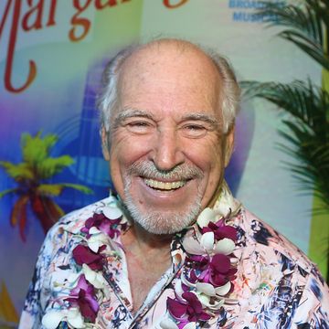 jimmy buffett smiles at the camera, he wears a pink hawaiian shirt with a purple and white lei