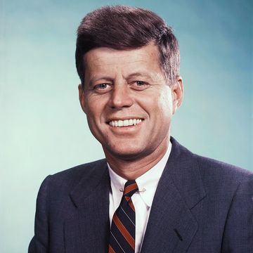 john f kennedy smiles at the camera, he wears a blue suit jacket, white collared shirt and blue and red striped tie