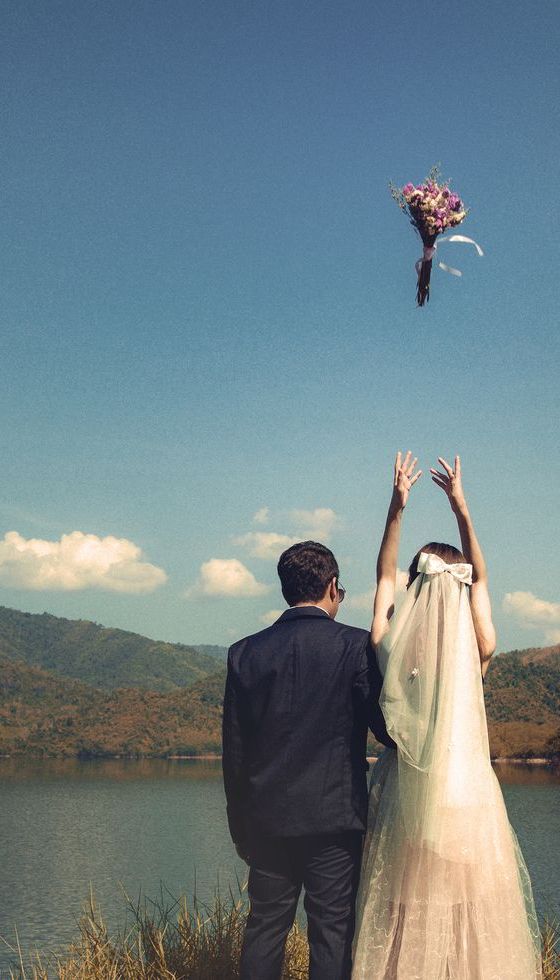 Couple Throwing Bouquet While Standing At Lakeshore Against Sky