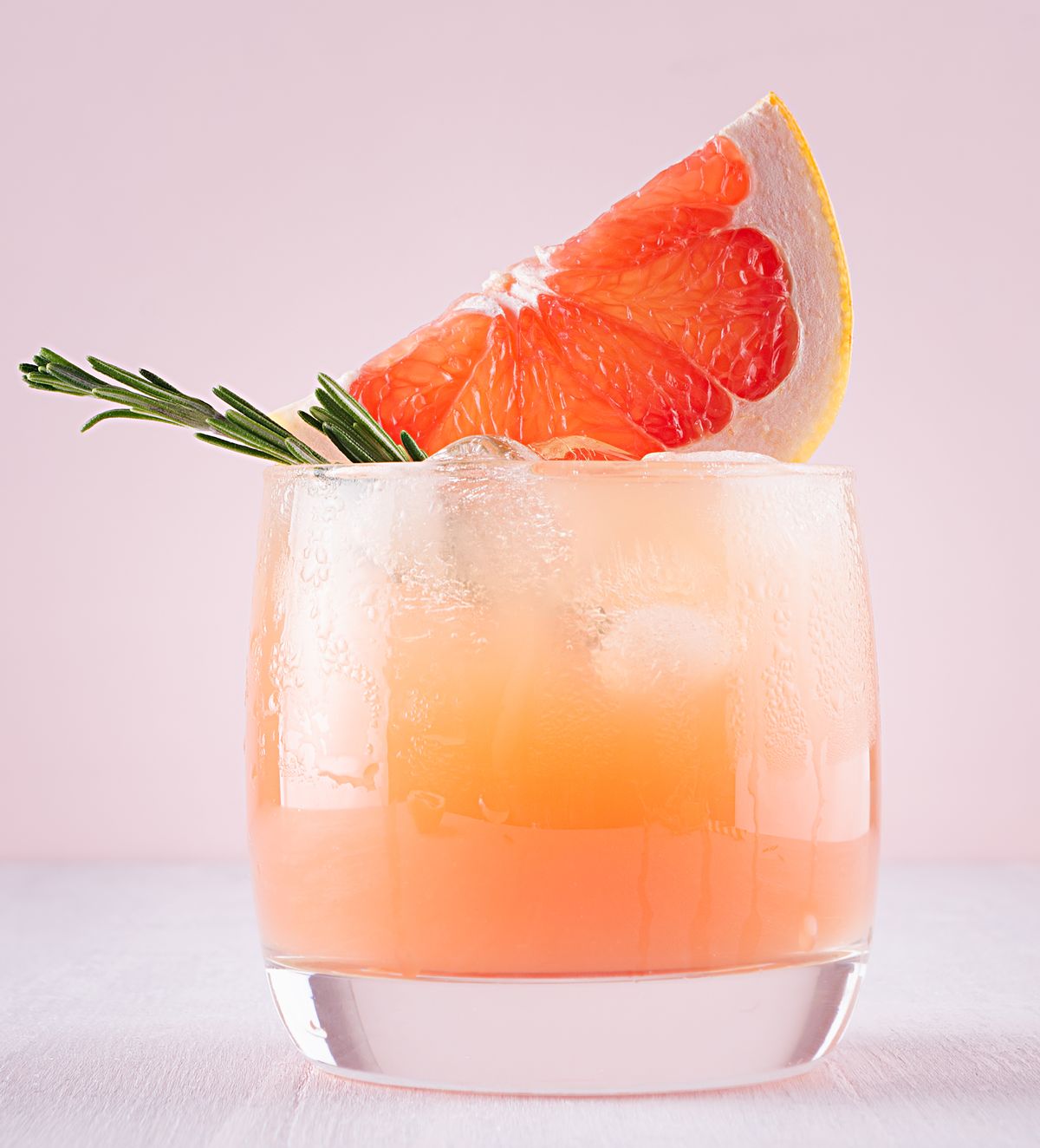 cold detox cocktail of grapefruit juice with ice, rosemary, slices citrus on soft light pink and white background