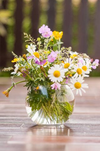 a colorful bouquet of freshly picked wild flowers from a natural meadow and garden presented on a table outdoors in a clear round bubble bowl glass vase fence in the background