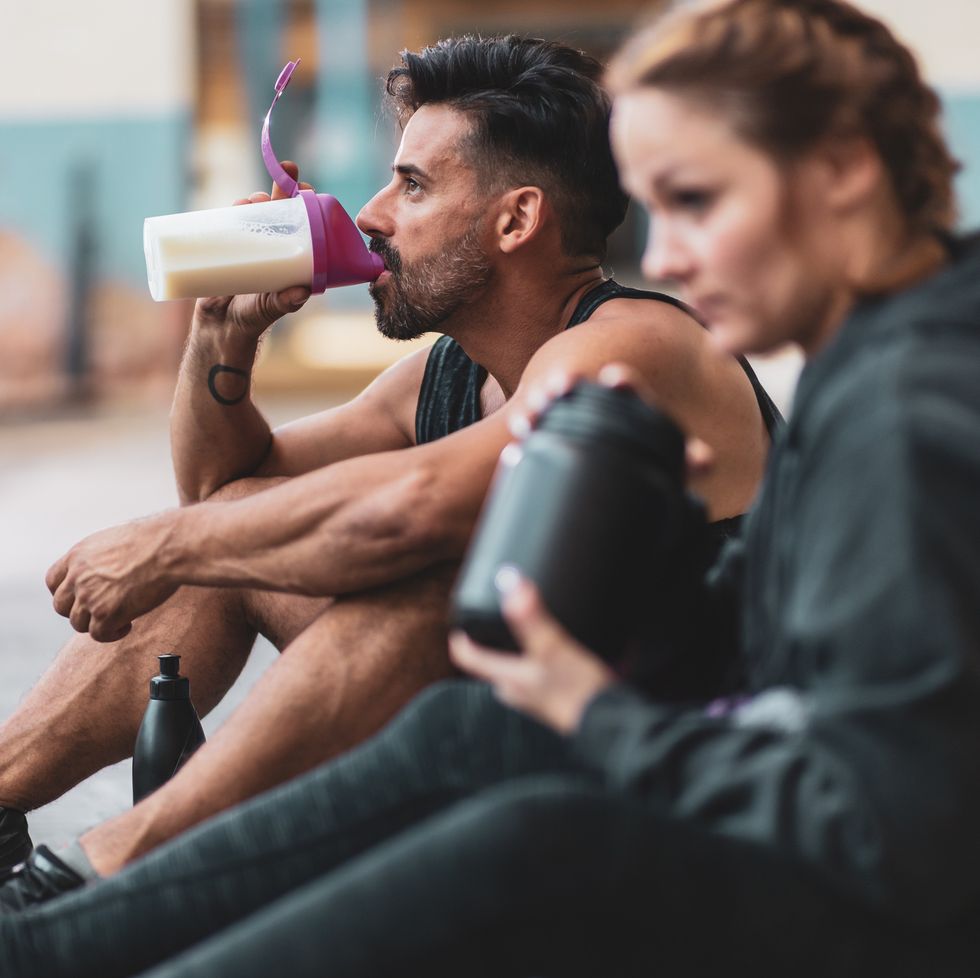 a man and a woman in sports clothing making and drinking protein drinks while sitting on the concrete floor