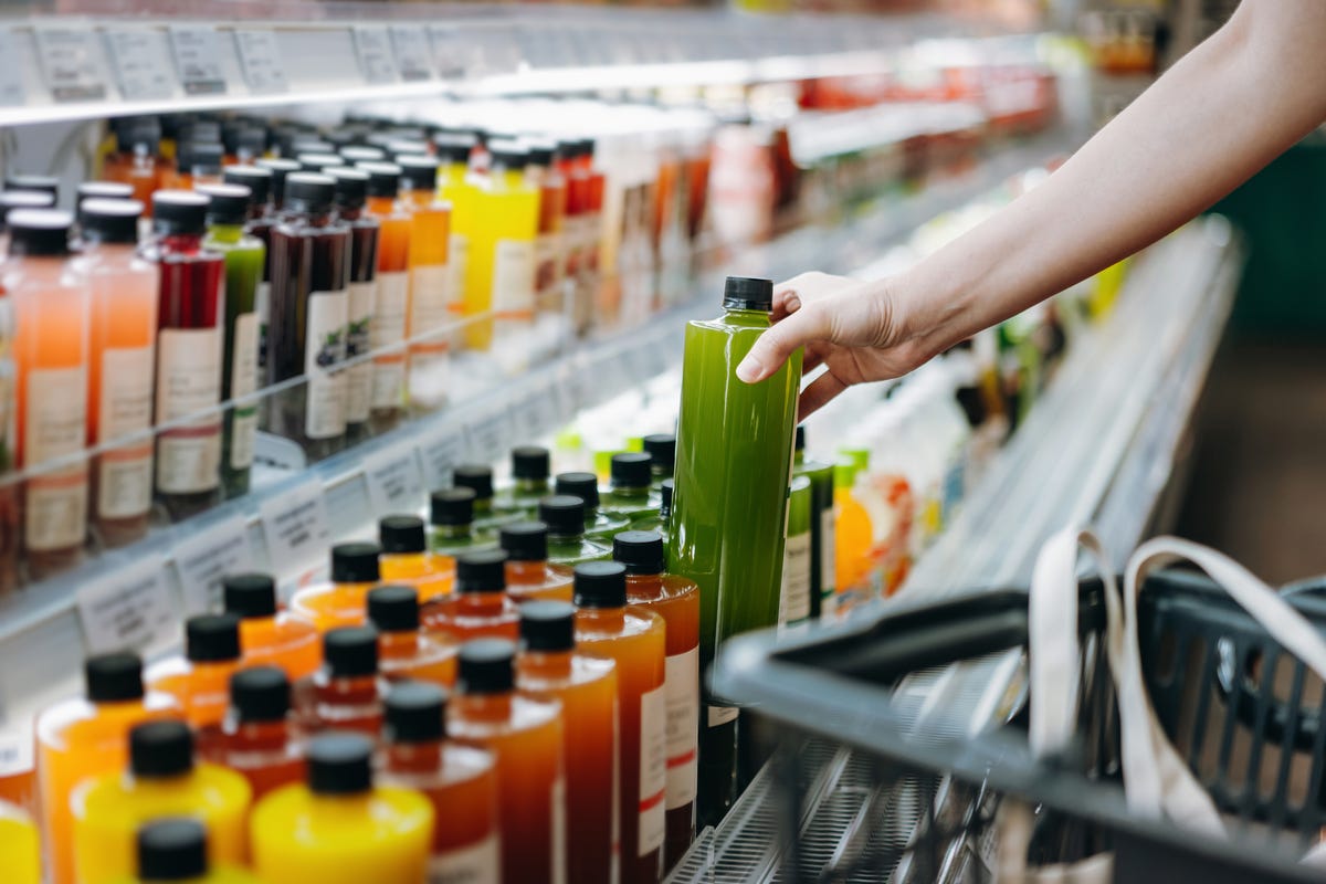 cropped shot of young asian woman shopping for fresh fruit juice from refrigerated shelves in supermarket and putting a bottle of fresh squeezed orange juice into cotton mesh eco bag in a shopping cart zero waste concept