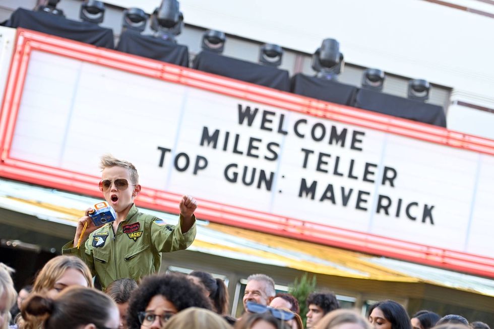 a boy in a flight suit in front of a marquee for top gun maverick maverick could be a popular baby boy name in 2023 fitting in with the neo cowboy name trend