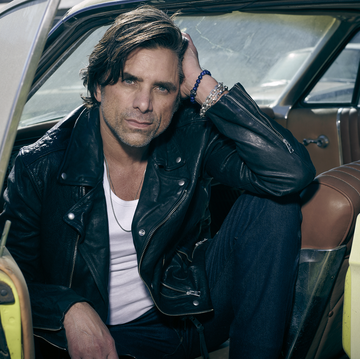 john stamos in a leather jacket and white tank top sitting in a old style car