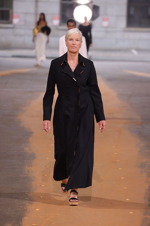 a white woman with white hair walks down a runway wearing a black coat