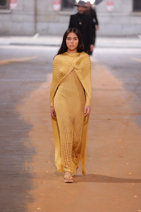 a brunette woman wearing a gold column dress walks down a runway