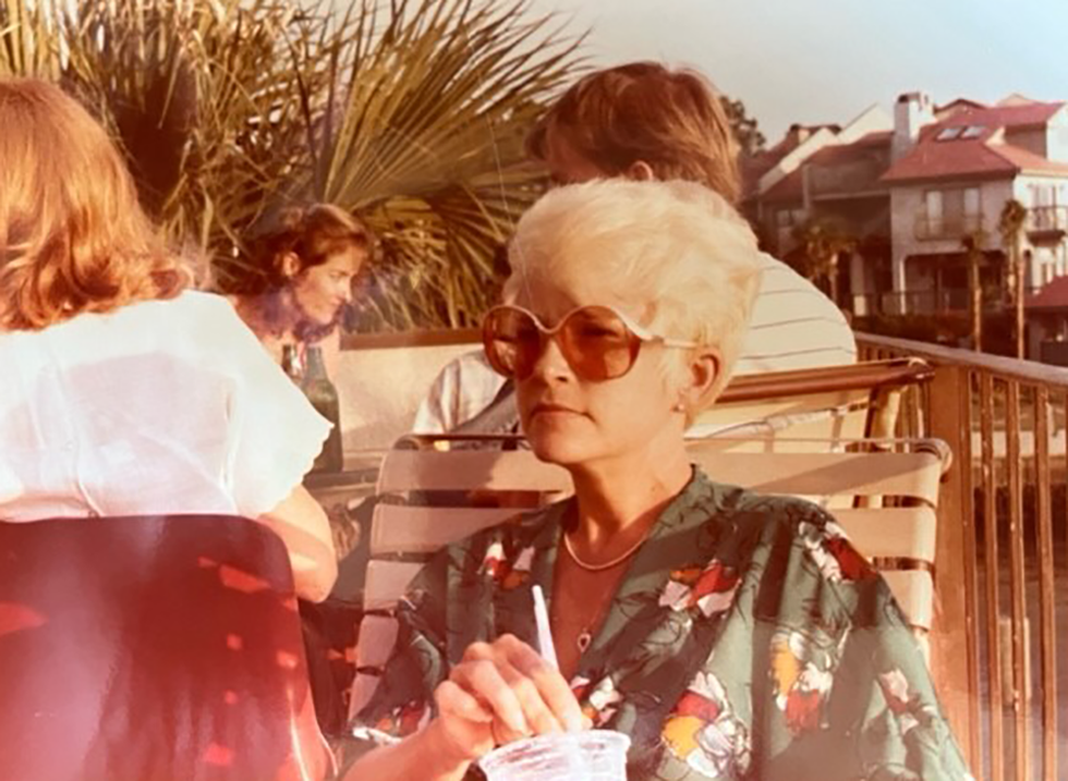 may cobbs mom a woman with short bright blonde hair wearing sunglasses and a floral shirt looking off into the distance holding a drink