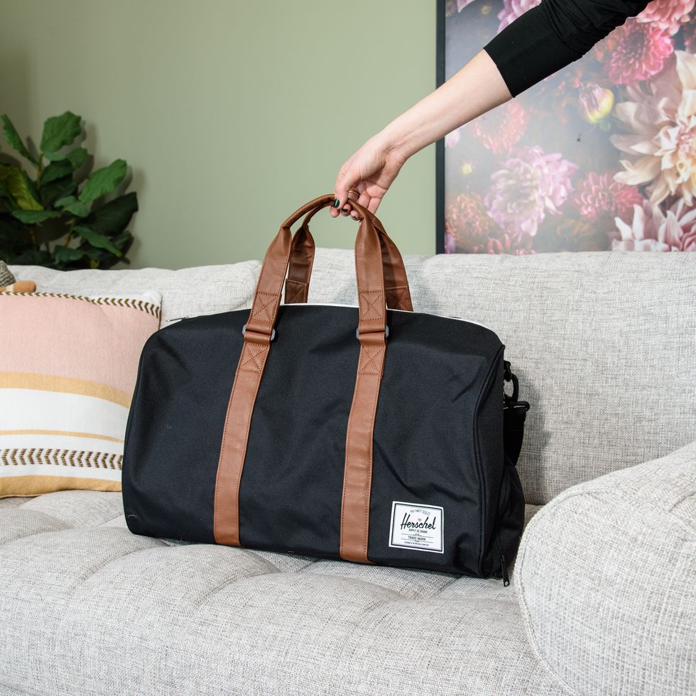 a black herschel novel duffel with brown leather straps on a grey couch, good housekeeping's best weekender bags