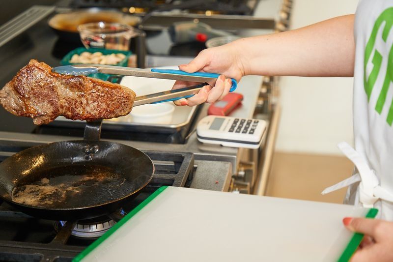 a person with tongs removing a browned steak from a carbon steel pan