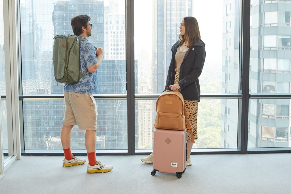 two people in front of windows one is carrying a large green backpack and another has a light tan backpack on top of a pink carry on luggage