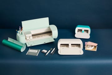 three cricut machines on a blue set with tools surrounding them, good housekeeping's best cricut machines
