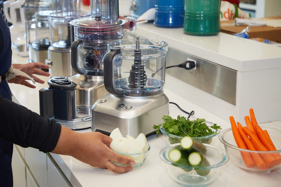 food processors on a white countertop with bowl of carrots, zucchini and onions